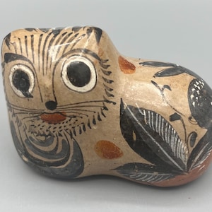 May include: Hand-painted ceramic figurine of a cat with black and white details, lying down with its head turned to the side. The cat has large eyes and a whimsical expression. The figurine is decorated with black and white floral patterns.