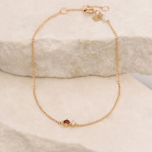Può includere: Delicato braccialetto con catena dorata con una piccola gemma rossa rotonda e una piccola gemma trasparente rotonda. Il braccialetto ha una chiusura a moschettone e un piccolo ciondolo a forma di cuore. Il braccialetto è esposto su una superficie bianca.