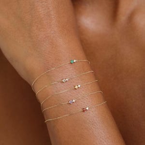 May include: Fine gold chain bracelets, each featuring a single gemstone. The gemstones are emerald green, clear, yellow, purple, and orange. The bracelets are shown on an arm.