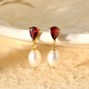 Puede incluir: Pendientes de oro con una piedra roja en forma de lágrima y una perla blanca que cuelga debajo.