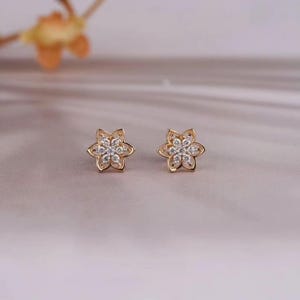 Könnte beinhalten: Ein Paar goldfarbene Ohrstecker. Jeder Ohrring hat ein Blumenmuster mit sechs Blütenblättern. Die Mitte jeder Blume ist mit kleinen, klaren Edelsteinen gefüllt. Die Ohrringe werden auf einer hellen Oberfläche präsentiert.