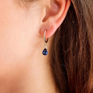 May include: A gold hoop earring featuring a small, clear, sparkling stone at the top and a dangling, pear-shaped, deep blue sapphire-coloured gemstone at the bottom. The hoop itself is adorned with a series of tiny, clear, pavé-set stones.