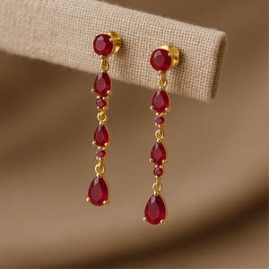 May include: Gold-coloured drop earrings with multiple red gemstones. The earrings feature a round gemstone at the top, followed by a series of smaller round and teardrop-shaped red gemstones, creating a dangling effect. The earrings are displayed against a neutral background.