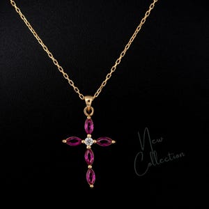 Pode incluir: Um colar dourado com um pingente em forma de cruz adornado com pedras rosa e um diamante central. A corrente é fina e o fundo é preto. As palavras "New Collection" estão no canto inferior direito.
