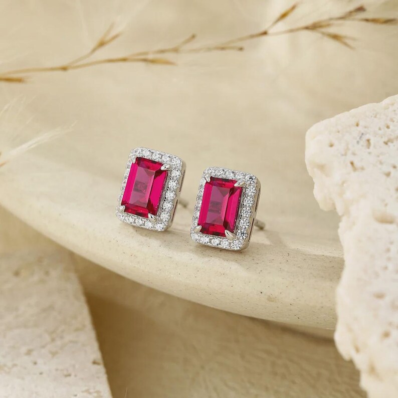 May include: A pair of stud earrings featuring rectangular emerald-cut vibrant red center stones. Each red stone is surrounded by a halo of small, clear, round-cut white stones set in a polished silver-toned metal frame with a secure post backing.
