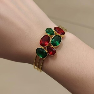 Può includere: Bracciale a polsino dorato con un gruppo di pietre preziose ovali rosse e verdi. Il bracciale ha un design a tre bande ed è indossato al polso. Le pietre preziose sono disposte in un motivo decorativo.