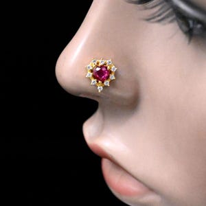 May include: A gold nose stud with a heart-shaped, deep red gemstone at its centre, encircled by smaller, clear gemstones. The stud is set against the skin, with a black background.