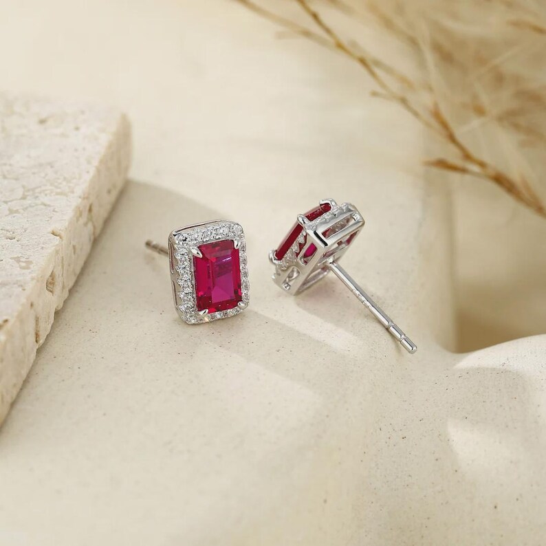 May include: A pair of stud earrings featuring rectangular, emerald-cut red gemstones set in silver-toned metal. Each central red stone is surrounded by a halo of small, clear, sparkling stones. The earrings include post-style backings for attachment.