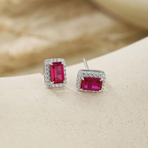 May include: A pair of stud earrings featuring rectangular, emerald-cut vibrant red center stones. Each central red stone is surrounded by a halo of small, clear, round-cut stones set in a polished silver-toned metal frame with a post backing.