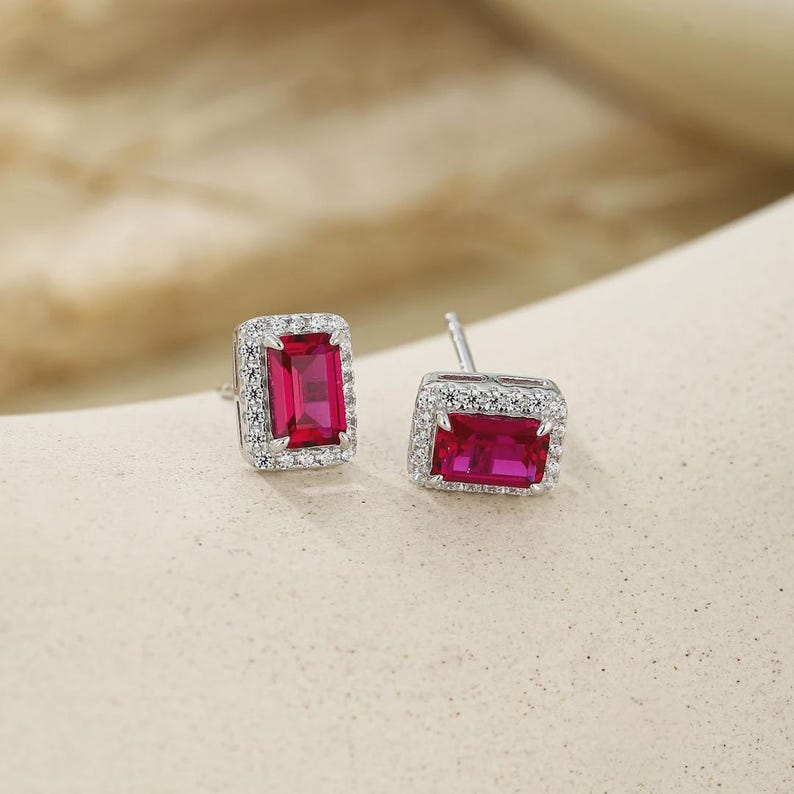 May include: A pair of stud earrings featuring rectangular, emerald-cut vibrant red center stones. Each central red stone is surrounded by a halo of small, clear, round-cut stones set in a polished silver-toned metal frame with a post backing.