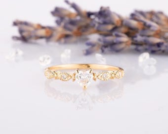May include: A delicate gold ring with a heart-shaped diamond and a band adorned with smaller diamonds. The ring is set against a white background with lavender flowers.