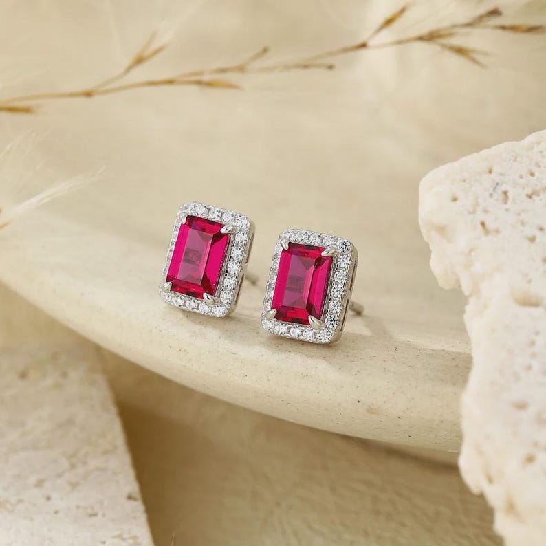 May include: A pair of stud earrings featuring rectangular emerald-cut vibrant red center stones. Each red stone is surrounded by a halo of small, clear, round-cut white stones set in a polished silver-toned metal frame with a secure post backing.