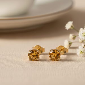 Puede incluir: Un par de pendientes de oro con gemas redondas de color ámbar. Los pendientes están engastados en un engaste de cuatro puntas y se muestran sobre una superficie de tono neutro, con un fondo borroso que incluye un plato blanco.