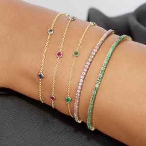 Puede incluir: Una colección de pulseras de oro adornan una muñeca. Las pulseras incluyen delicadas cadenas con piedras preciosas de colores y pulseras de tenis con piedras rosas y verdes. Las piedras preciosas incluyen piedras azules, rojas y verdes.