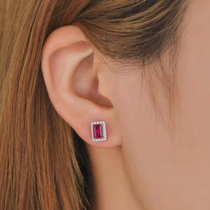 May include: A rectangular stud earring featuring a vibrant fuchsia-red center stone surrounded by a halo of small, clear, sparkling stones set in a polished silver-toned metal frame, securely fastened to the earlobe.