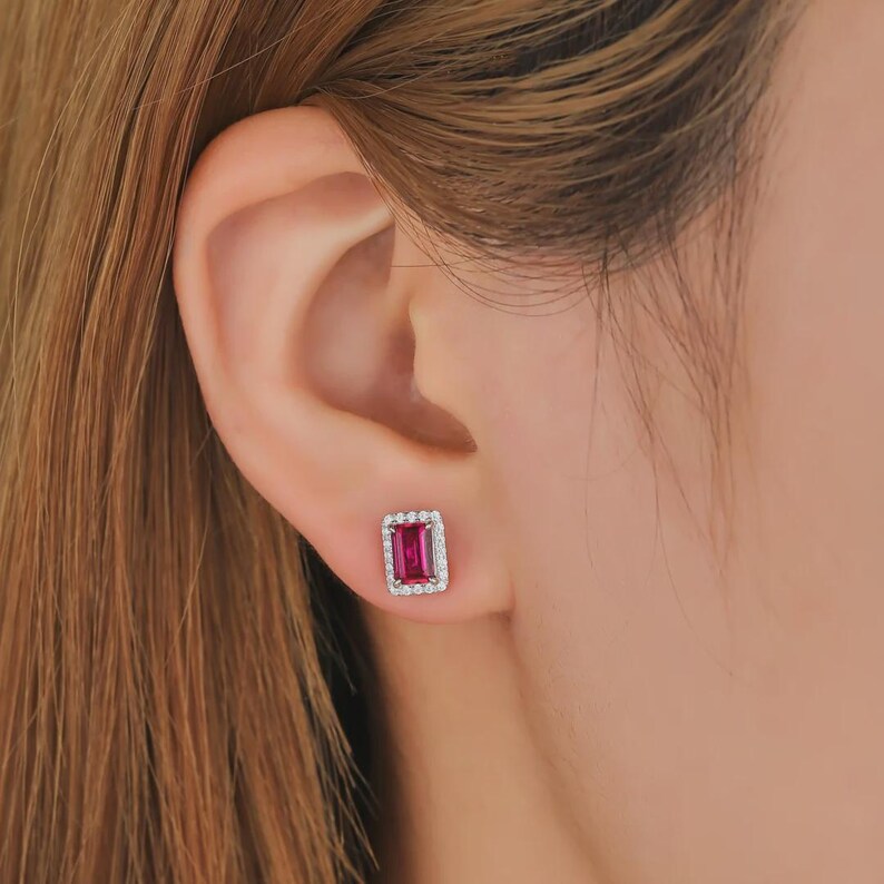 May include: A rectangular stud earring featuring a vibrant fuchsia-red center stone surrounded by a halo of small, clear, sparkling stones set in a polished silver-toned metal frame, securely fastened to the earlobe.