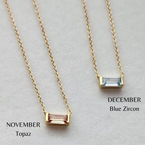 May include: Two gold necklaces with rectangular gemstones. One features a pink topaz, labeled "November Topaz," and the other a blue zircon, labeled "December Blue Zircon." The necklaces have delicate gold chains.