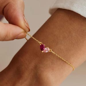 Könnte beinhalten: Ein filigranes goldenes Armband mit zwei tropfenförmigen Edelsteinen, einem rubinroten und einem hellrosa. Das Armband wird von einer Hand gehalten, wodurch die feine Kette und die leuchtenden Farben der Steine hervorgehoben werden.