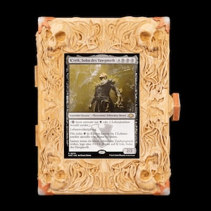 May include: A rectangular light-colored wooden frame with intricate carvings of skulls and swirling organic patterns at each corner. The frame holds a Magic: The Gathering trading card titled K'rrik, Sohn des Yawgmoth, featuring a dark figure.