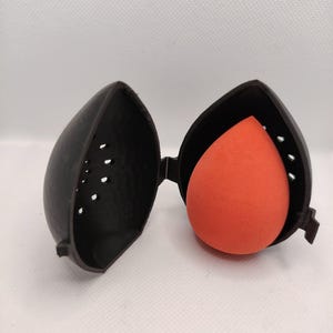 May include: A black, 3D-printed case, open to reveal an orange makeup sponge. The case is shaped like a stylized teardrop with small ventilation holes. The sponge is a vibrant orange color and is teardrop-shaped.