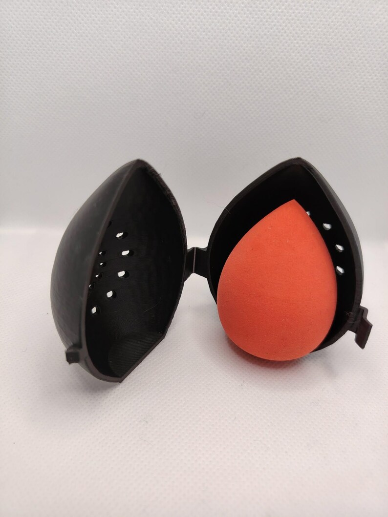 May include: A black, 3D-printed case, open to reveal an orange makeup sponge. The case is shaped like a stylized teardrop with small ventilation holes. The sponge is a vibrant orange color and is teardrop-shaped.