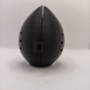 May include: A black, egg-shaped object with a vertical slit down the center and small circular holes on the sides. A small square indentation is visible near the bottom. The object appears to be made of a textured material.