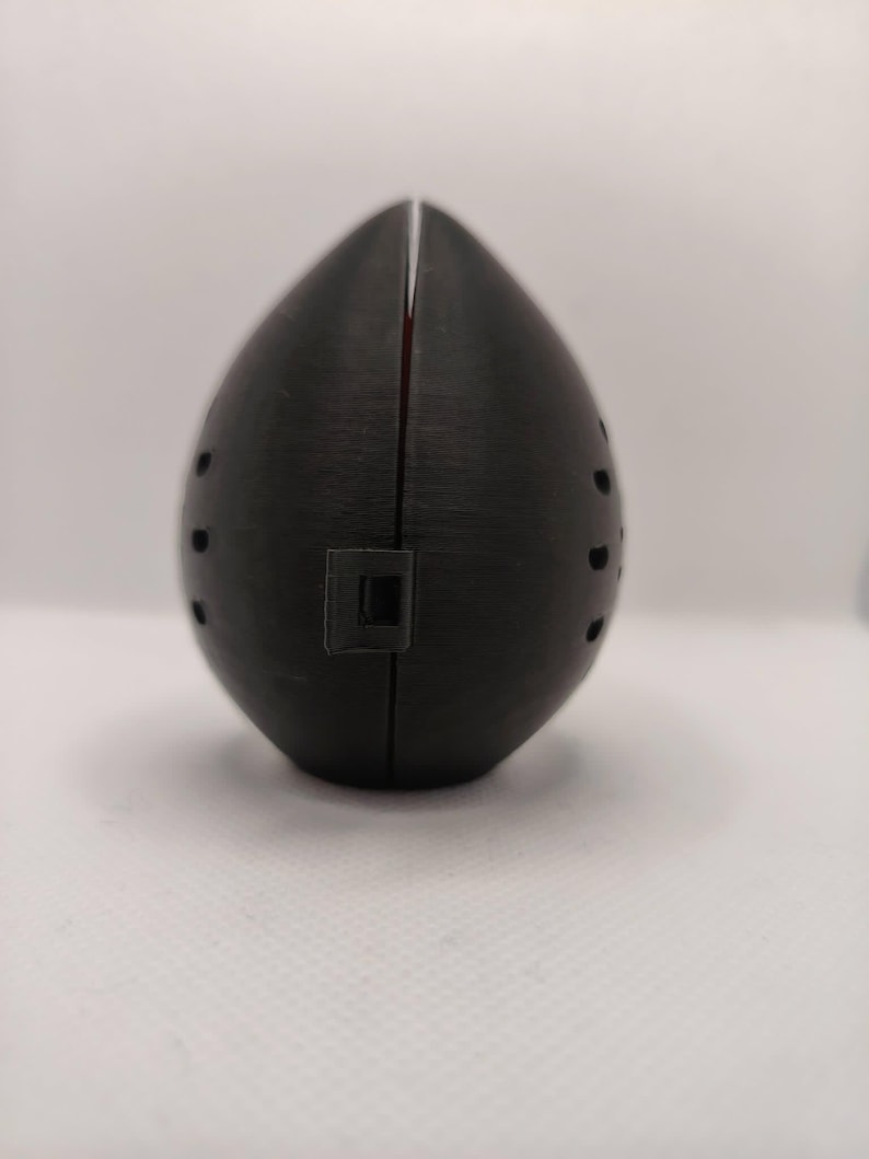 May include: A black, egg-shaped object with a vertical slit down the center and small circular holes on the sides. A small square indentation is visible near the bottom. The object appears to be made of a textured material.