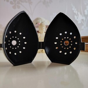 May include: Black, teardrop-shaped decorative object with a series of small, evenly spaced holes. The object is open, revealing the interior. The design is symmetrical, with a central circular pattern of holes. The object is sitting on a light-colored surface.