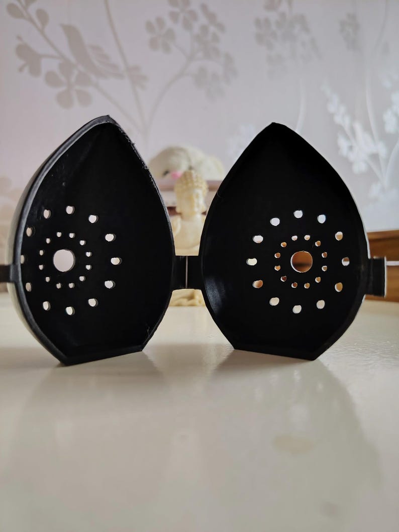 May include: Black, teardrop-shaped decorative object with a series of small, evenly spaced holes. The object is open, revealing the interior. The design is symmetrical, with a central circular pattern of holes. The object is sitting on a light-colored surface.