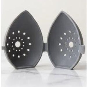 May include: A gray, hinged, teardrop-shaped container, open to reveal the interior. Each half has a central hole surrounded by smaller, evenly spaced perforations. The container is designed to hold and protect an item.