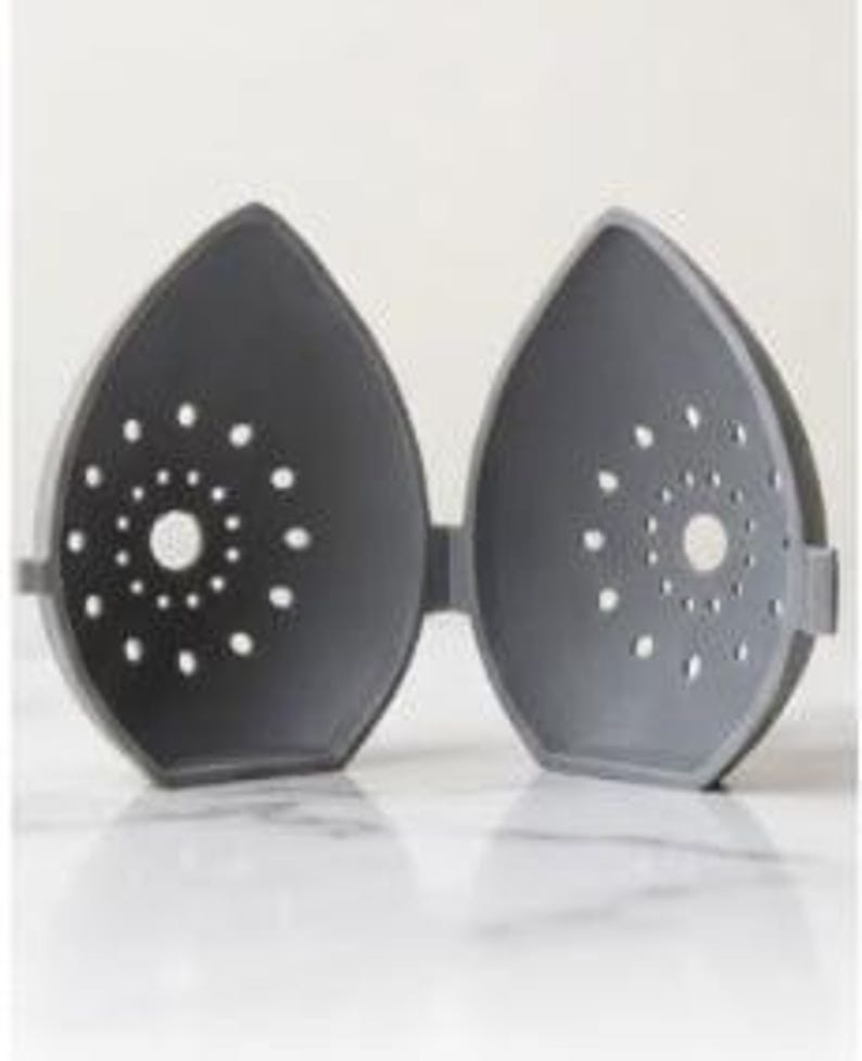 May include: A gray, hinged, teardrop-shaped container, open to reveal the interior. Each half has a central hole surrounded by smaller, evenly spaced perforations. The container is designed to hold and protect an item.