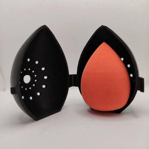 Può includere: Un astuccio nero a forma di goccia, aperto per rivelare una spugnetta per il trucco arancione. L'astuccio ha una serie di piccole perforazioni circolari su un lato. L'astuccio è incernierato e sembra essere di plastica.