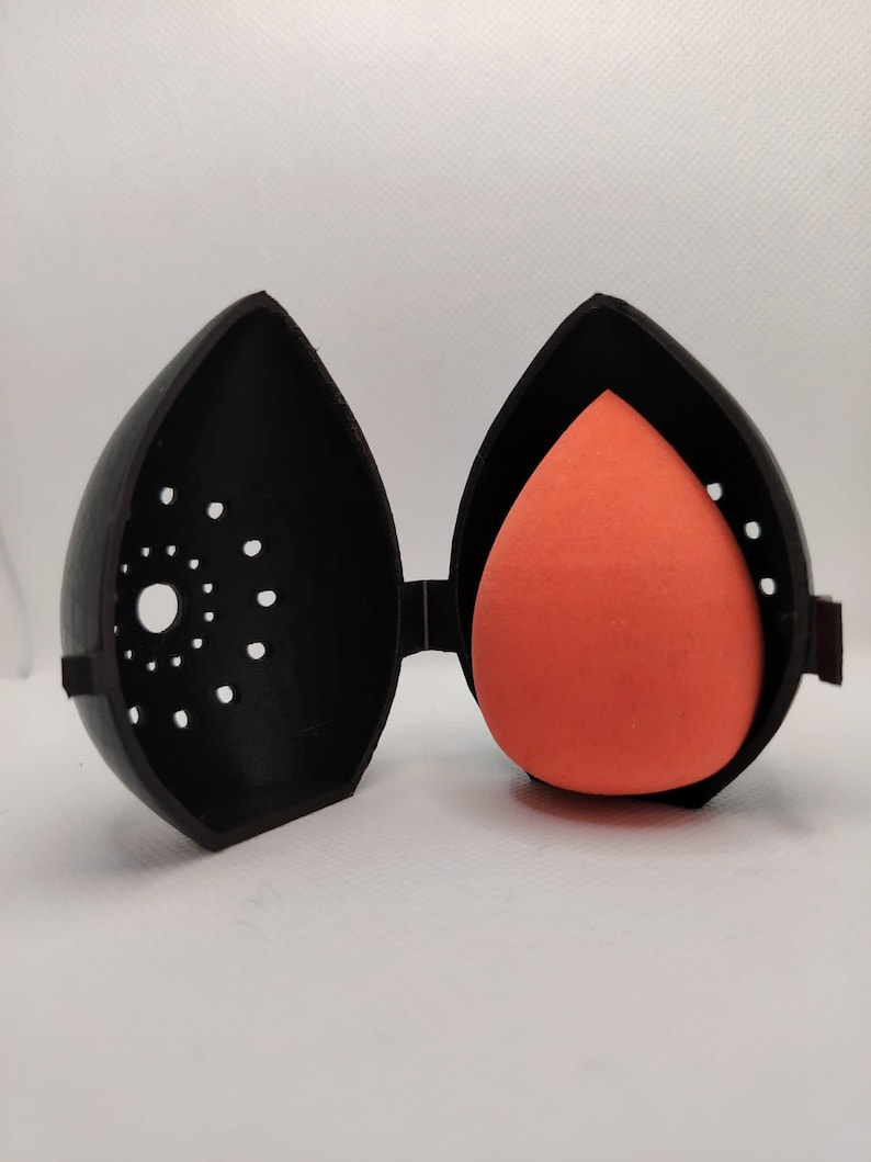 May include: A black, teardrop-shaped case, open to reveal an orange makeup sponge. The case has a series of small, circular perforations on one side. The case is hinged and appears to be made of plastic.