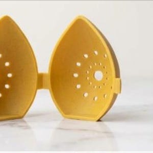 May include: A yellow, egg-shaped kitchen gadget, open to reveal two halves connected by a hinge. Each half has a series of small, circular holes and a larger central hole. The object is made of a matte material and sits on a white surface.