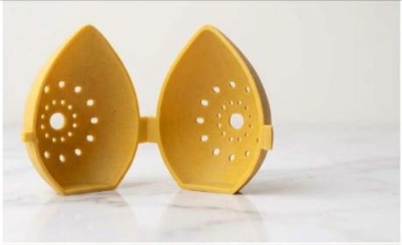 May include: A yellow, egg-shaped kitchen gadget, open to reveal two halves connected by a hinge. Each half has a series of small, circular holes and a larger central hole. The object is made of a matte material and sits on a white surface.