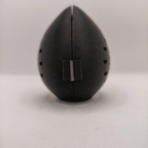 May include: A black, teardrop-shaped object with a vertical seam and small, evenly spaced holes. A rectangular silver-colored piece is attached near the bottom. The object is set against a plain white background.