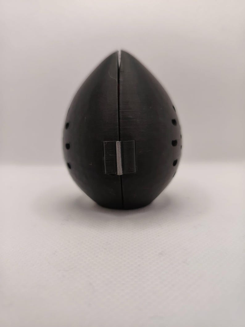 May include: A black, teardrop-shaped object with a vertical seam and small, evenly spaced holes. A rectangular silver-colored piece is attached near the bottom. The object is set against a plain white background.