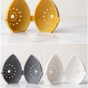 May include: A set of three egg-shaped kitchen gadgets in yellow, gray, and white. Each has a hinged design and a series of small, circular holes. The yellow one is open, while the others are closed. The gadgets are on a white marble surface.
