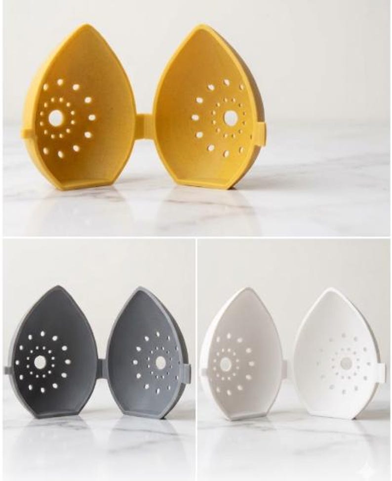 May include: A set of three egg-shaped kitchen gadgets in yellow, gray, and white. Each has a hinged design and a series of small, circular holes. The yellow one is open, while the others are closed. The gadgets are on a white marble surface.