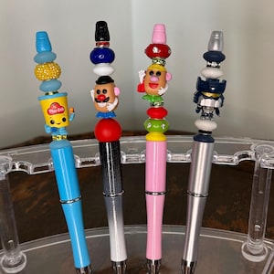 May include: Four decorative pens with colorful, beaded toppers. The pens have blue, black and white, pink, and silver barrels. The toppers feature various designs, including a Play-Doh character, a Mr. Potato Head, and a robot.