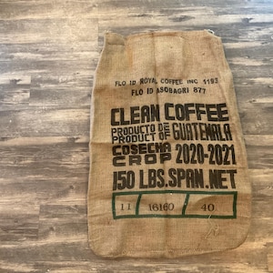 May include: A brown burlap sack with black text that reads "Clean Coffee", "Producto de Guatemala", "Cosecha Crop 2020-2021", and "150 LBS.SPAN.NET". The sack also has the text "FLO ID ROYAL COFFEE INC 1193" and "FLO ID ASOBAGRI 877".