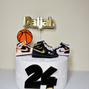 May include: A white cake with black and gold decorations. The cake is decorated with a basketball, two black and white sneakers, and the name "Daijah" in gold letters. The number "24" is also on the cake in black with gold accents.