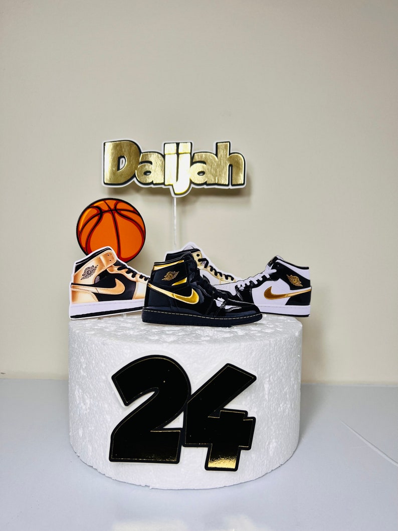 May include: A white cake with black and gold decorations. The cake is decorated with a basketball, two black and white sneakers, and the name "Daijah" in gold letters. The number "24" is also on the cake in black with gold accents.