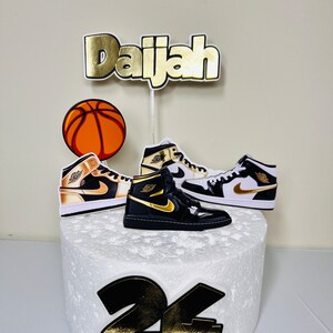 May include: A white cake with gold and black decorations. The cake is decorated with a basketball, two sneakers, and the name "Daijah" in gold letters. The number "24" is also on the cake in gold and black.