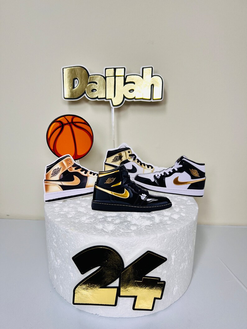May include: A white cake with gold and black decorations. The cake is decorated with a basketball, two sneakers, and the name "Daijah" in gold letters. The number "24" is also on the cake in gold and black.
