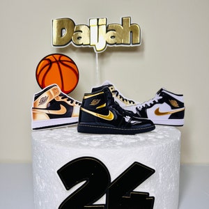 May include: A white cake decorated with a basketball, two black and gold sneakers, and the name "Daijah" in gold letters. The number "24" is also on the cake in black and gold.