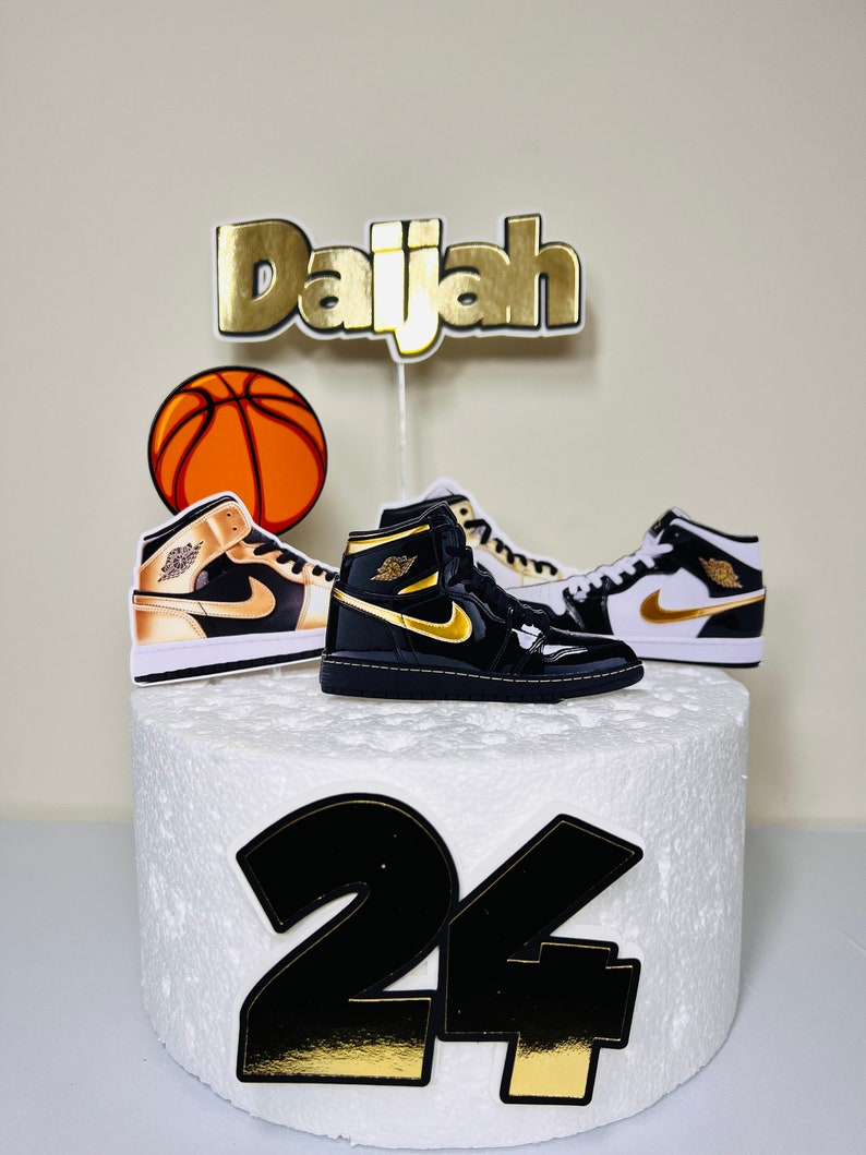 May include: A white cake decorated with a basketball, two black and gold sneakers, and the name "Daijah" in gold letters. The number "24" is also on the cake in black and gold.
