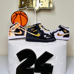May include: A white cake decorated with a basketball, two black and gold sneakers, and the name "Daijah" in gold letters. The number "24" is also on the cake in black with gold trim.