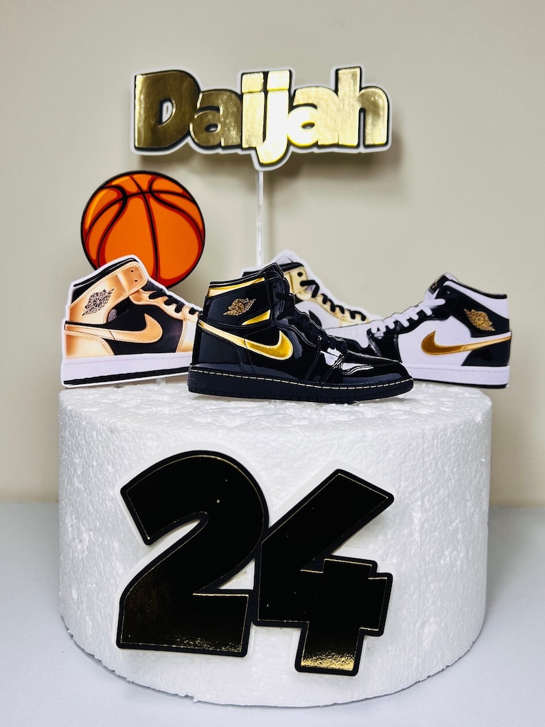 May include: A white cake decorated with a basketball, two black and gold sneakers, and the name "Daijah" in gold letters. The number "24" is also on the cake in black with gold trim.