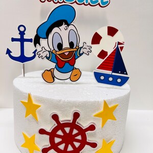 May include: A white cake decorated with a blue and yellow nautical themed cake topper. The topper features a cartoon duck, an anchor, a life preserver, a sailboat, and a red steering wheel. The cake is decorated with yellow stars. The cake topper says "Miguel".